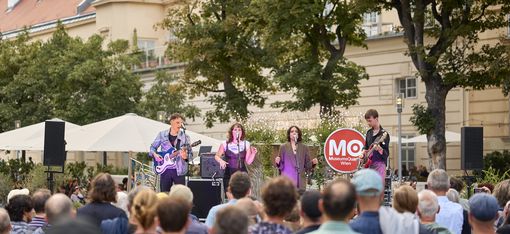 Mehrere Musiker mit Instrumenten und Mikrofonen stehen auf einer Bühne vor einer Menschenmenge im Freien, im Hintergrund Bäume und ein Gebäude mit roten Dachziegeln.