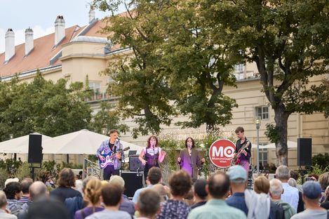 Mehrere Musiker mit Instrumenten und Mikrofonen stehen auf einer Bühne vor einer Menschenmenge im Freien, im Hintergrund Bäume und ein Gebäude mit roten Dachziegeln.