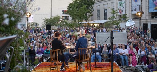 Two people sitting on chairs on an orange carpet facing a large outdoor audience between buildings.