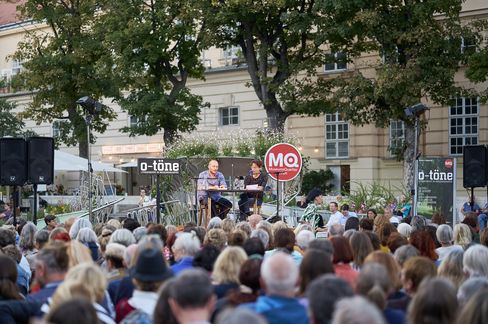 Man sitting at a table on a stage speaking into a microphone to an outdoor audience in front of a historic building