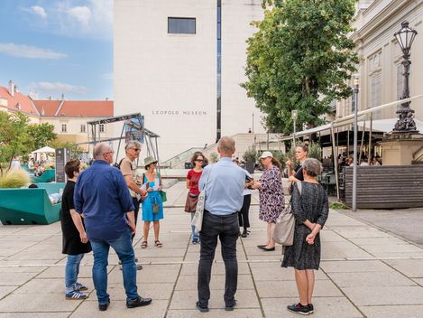 Eine Gruppe von Personen steht im MQ Innenhof und hört einer Führung zu.