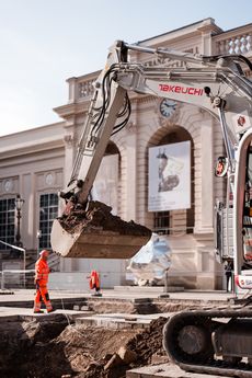 Bagger mit ausgehobener Schaufel vor einem historischen Gebäude und zwei Bauarbeitern in orangefarbener Schutzkleidung