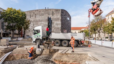 Baustelle mit zwei Bauarbeitern in orangener Schutzkleidung am MQ Hauptplatz, einem Lkw mit Kran und einem runden, mumok im Hintergrund