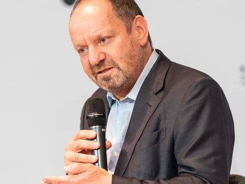 Sitzender Mann im Anzug hält ein Mikrofon und gestikuliert mit der freien Hand