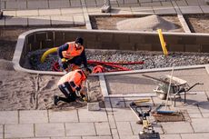 Zwei Bauarbeiter in orangen Warnwesten knien auf einem mit Kies bedeckten Boden und arbeiten an einem Kabel oder Rohr