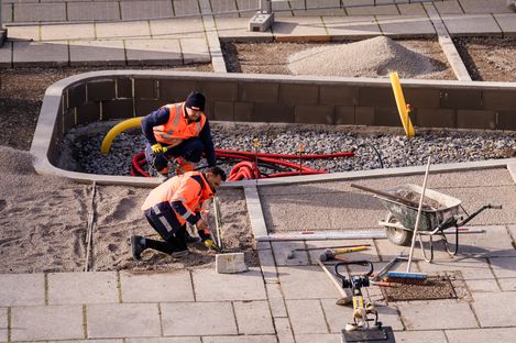 Zwei Bauarbeiter in orangen Warnwesten knien auf einem mit Kies bedeckten Boden und arbeiten an einem Kabel oder Rohr