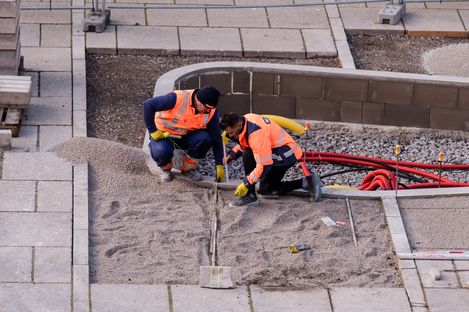Zwei Bauarbeiter in orangen Warnwesten knien auf einem mit Kies bedeckten Boden und arbeiten an einem Kabel oder Rohr