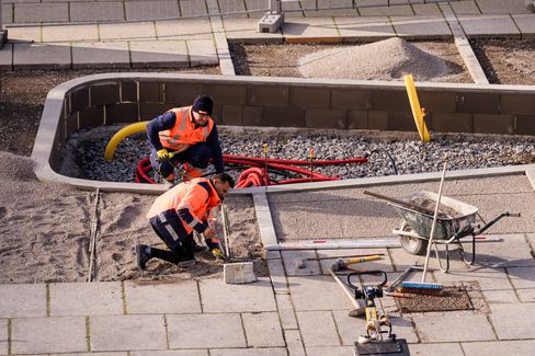 Zwei Bauarbeiter in orangen Warnwesten knien auf einem mit Kies bedeckten Boden und arbeiten an einem Kabel oder Rohr
