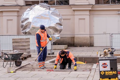Zwei Bauarbeiter mit orangen Warnwesten arbeiten auf einer Baustelle vor einem Gebäude mit einer großen silbernen Skulptur