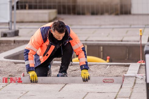 Ein Mann in orangener Warnweste, der etwas am Boden einer Baustelle abmisst.