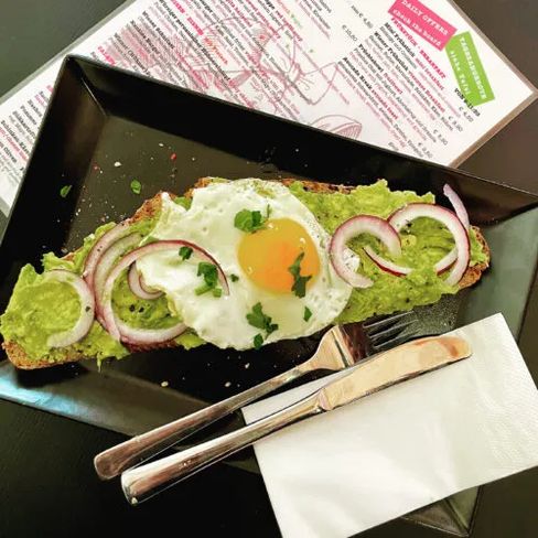 Slice of bread with avocado cream, fried egg, and red onion rings on a rectangular plate next to cutlery and menu.