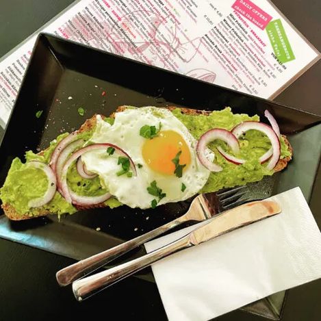 Slice of bread with avocado cream, fried egg, and red onion rings on a rectangular plate next to cutlery and menu.