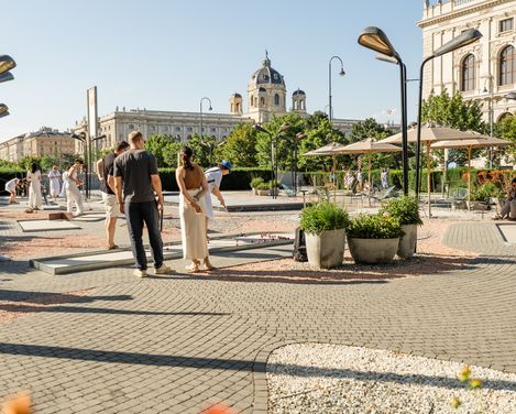 Menschen spielen Minigolf am Vorplatz des MQ. Im Hintergrund sind historische Gebäude und ein strahlend blauer Himmel zu sehen, was eine lebendige, entspannte Atmosphäre schafft.