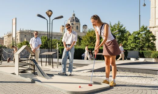 Several people play mini golf at a temporary course in MuseumsQuartier Vienna, surrounded by green areas.
