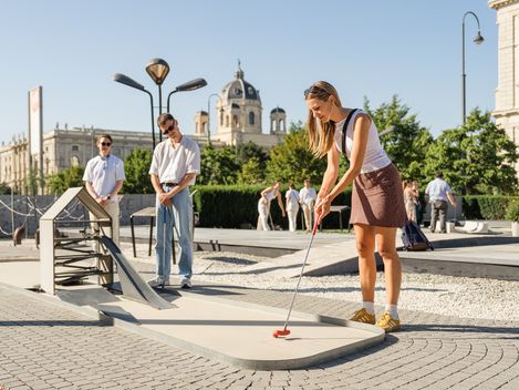 Drei Personen spielen Minigolf am MQ Amore am Vorplatz des MQ.