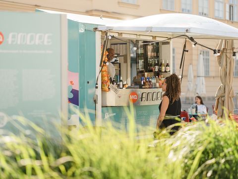 Frau mit lockigen Haaren steht vor einem türkisfarbenen Imbisswagen mit Getränken und Snacks, im Hintergrund ein helles Gebäude.