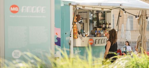 Woman with curly hair stands in front of a turquoise food truck with drinks and snacks, light-colored building in the background.