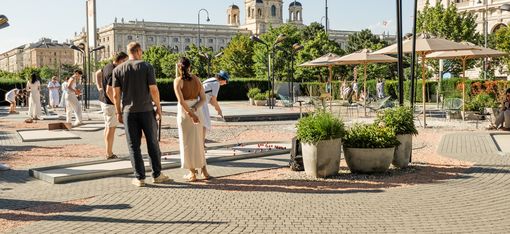 Menschen spielen Minigolf am Vorplatz des MQ. Im Hintergrund sind historische Gebäude und ein strahlend blauer Himmel zu sehen, was eine lebendige, entspannte Atmosphäre schafft.
