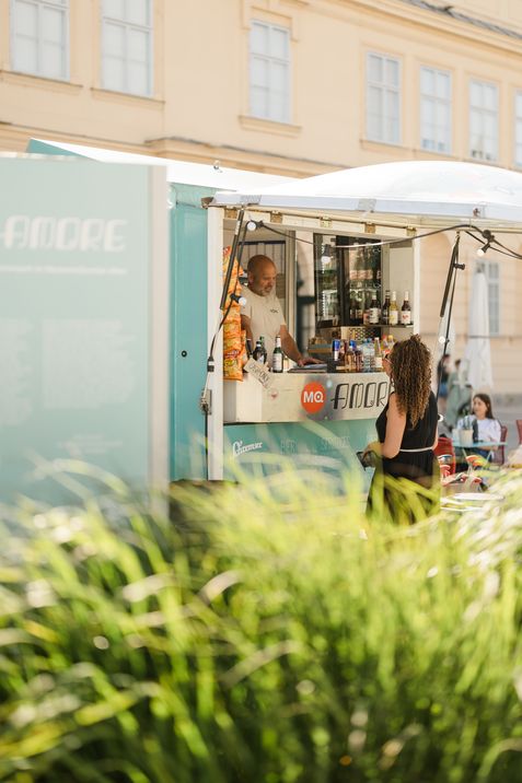 Woman with curly hair stands in front of a turquoise food truck with drinks and snacks, light-colored building in the background.