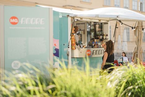 Woman with curly hair stands in front of a turquoise food truck with drinks and snacks, light-colored building in the background.