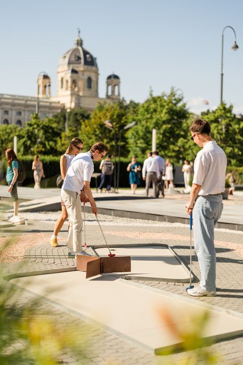 A person takes a mini golf shot at MuseumsQuartier Vienna while other visitors watch.