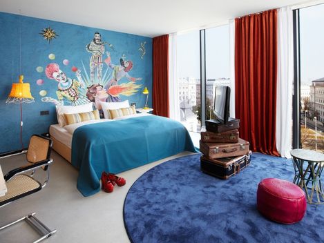 Lebhaftes Hotelzimmer mit einer blauen Wandtapete mit bunten Clowns, einem Bett mit blauer Bettwäsche und einem Stapel Koffer als TV-Ständer. Orangefarbene Vorhänge sorgen für Wärme.