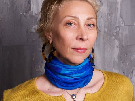 Oberkörper einer Person mit kurzem Haar, die ein senfgelbes Oberteil, einen blauen Schal und eine Halskette trägt.