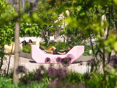 Person liegt auf einer modernen, hellen Liege im Freien, umgeben von Bäumen und Pflanzen in einem urbanen Park