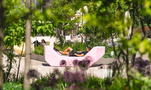 Person lying on a modern light-colored lounge chair outdoors surrounded by trees and plants in an urban park