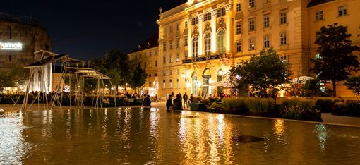 Nachtansicht eines beleuchteten Gebäudes im europäischen Stil, das sich in einem ruhigen Teich spiegelt, mit Menschen, die sich darum versammelt haben, und einer modernen Skulptur in der Nähe; ruhige Atmosphäre.