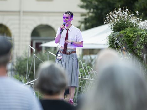 Person wearing a tie speaking into a microphone on an outdoor stage in front of an audience.