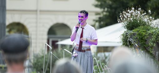 Person wearing a tie speaking into a microphone on an outdoor stage in front of an audience.