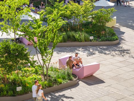 Zwei Personen sitzen auf einer pinkfarbenen, geometrisch geformten Bank vor einem bepflanzten Bereich mit Bäumen und Sträuchern in einem Innenhof mit Cafés im Hintergrund