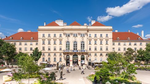 Blick auf den Haupteingang des MuseumsQuartier Wien mit historischer Fassade, begrünten Aufenthaltsbereichen und Besucher:innen im Innenhof bei blauem Himmel.