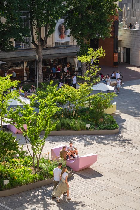 Städtischer Platz mit bepflanzten Beeten, rosa Sitzbänken und Menschen, die sich im Schatten von Bäumen aufhalten