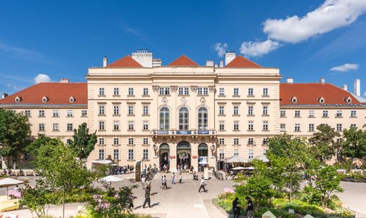 Blick auf den Haupteingang des MuseumsQuartier Wien mit historischer Fassade, begrünten Aufenthaltsbereichen und Besucher:innen im Innenhof bei blauem Himmel.