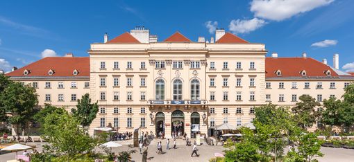 Blick über den begrünten Haupthof des MuseumsQuartier Wien auf das historische Haupteingangsgebäude. Besucher:innen bewegen sich zwischen Bäumen, Blumenbeeten, Sitzmöbeln und Gastgärten unter blauem Himmel.