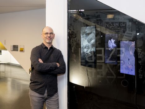 Man with glasses and dark shirt leaning with crossed arms against a white wall next to a glass panel showing abstract images in a modern interior
