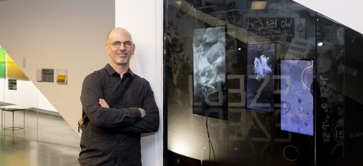 Mann mit Brille und dunklem Hemd lehnt mit verschränkten Armen an einer weißen Wand neben einer Glasfläche mit abstrakten Bildern im Hintergrund eines modernen Innenraums