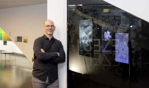 Mann mit Brille und dunklem Hemd lehnt mit verschränkten Armen an einer weißen Wand neben einer Glasfläche mit abstrakten Bildern im Hintergrund eines modernen Innenraums