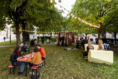 People sitting at a red table and on benches under trees with string lights, a kiosk and additional seating visible on a grassy park area in the background.