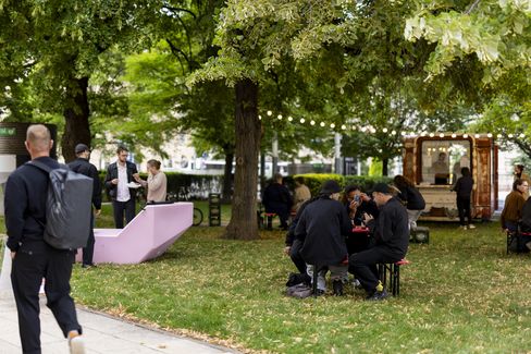 People sitting at tables and benches under trees with string lights in a park next to a kiosk.