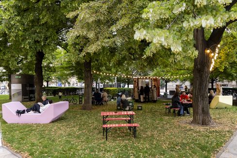 Park with trees, benches, and people sitting and relaxing near a small kiosk with string lights