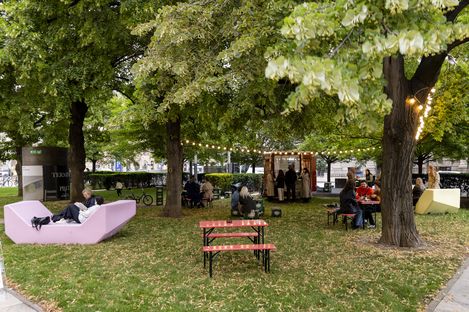 Park with trees, benches, and people sitting and relaxing near a small kiosk with string lights
