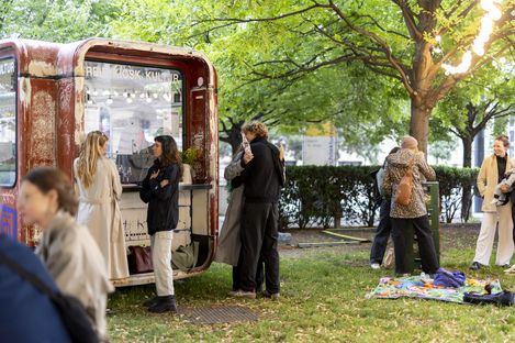 Several people stand and talk in front of a small red kiosk in a park with trees and grass