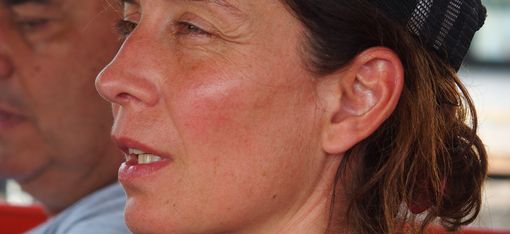 Nahaufnahme einer Frau mit dunklem Netzcap, die nach links blickt, im Hintergrund unscharf ein Mann