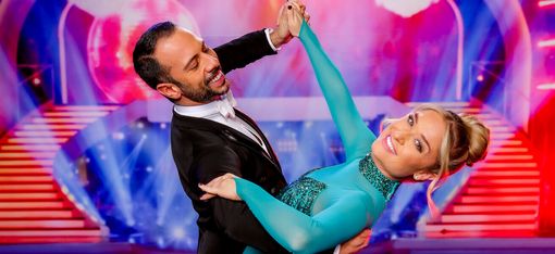 Ein Paar in eleganter Tanzpose auf einer Bühne mit buntem Hintergrund und Treppen