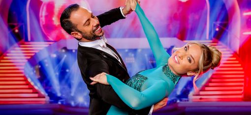Ein Paar in eleganter Tanzpose auf einer Bühne mit buntem Hintergrund und Treppen