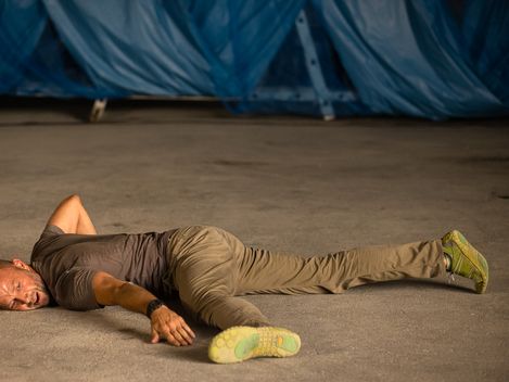 Mann liegt ausgestreckt auf dem Boden mit einem Arm über dem Kopf und trägt ein T-Shirt, Hose und Sportschuhe