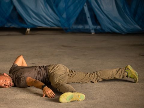 Man lying stretched out on the ground with one arm over his head wearing a t-shirt, pants, and athletic shoes